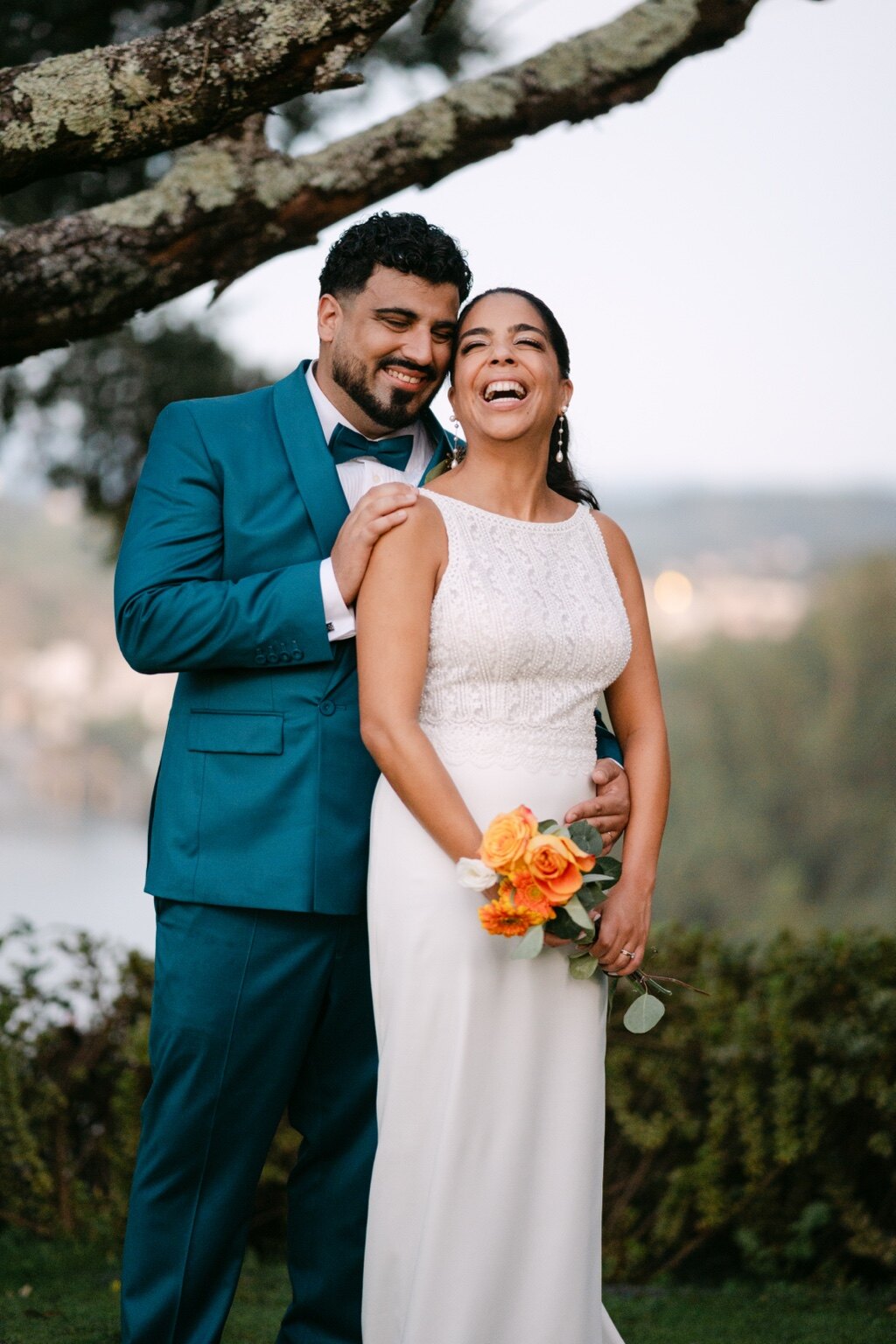 Um casal ri num retrato espontâneo ao ar livre, junto a uma árvore.