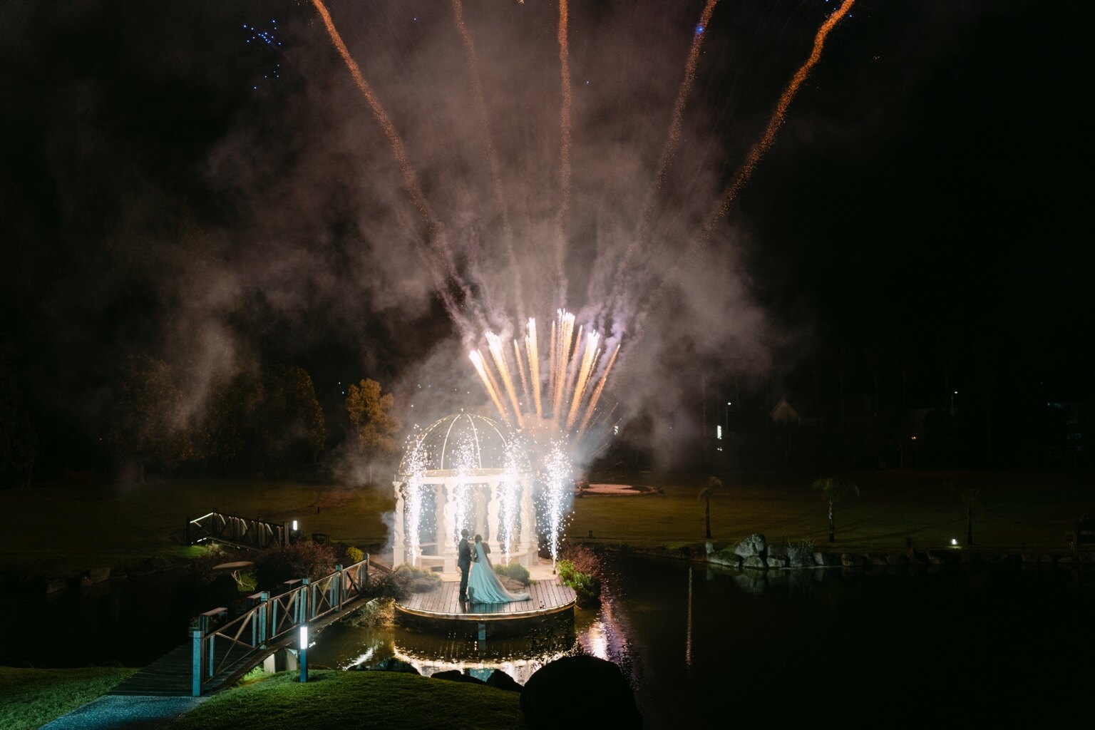 Fogo de artifício ilumina a noite sobre um coreto junto à água.